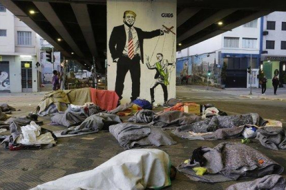 Un grupo de sin techo duerme bajo un puente de Sao Paulo ante una pintada de Bolsonaro como marioneta de Trump.-NELSON ANTOINE (AP)