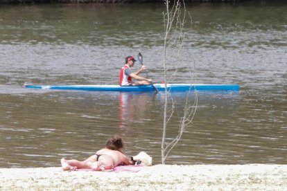 Una mujer toma el sol en la arena ante el paso de una piragüa por el río.- J. M. LOSTAU