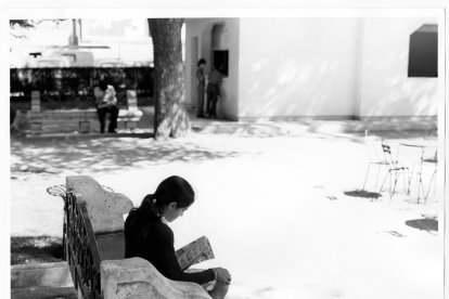 Una niña lee sentada en un banco de plaza Poniente, al fondo se ve el edificio de una de las bibliotecas de verano