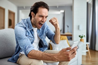 Happy man reading good news over smart phone and celebrating at home.