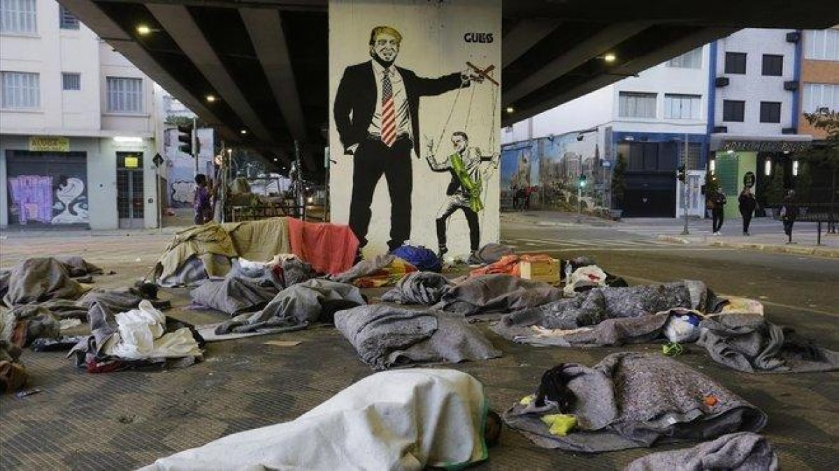 Un grupo de sin techo duerme bajo un puente de Sao Paulo ante una pintada de Bolsonaro como marioneta de Trump.-NELSON ANTOINE (AP)