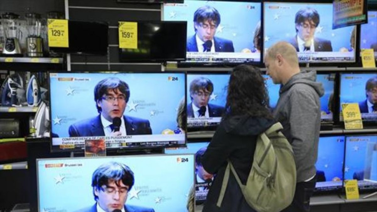 Una pareja sigue la rueda de prensa de Puigdemont en una tienda del Poblenou.-/ FERRAN NADEU